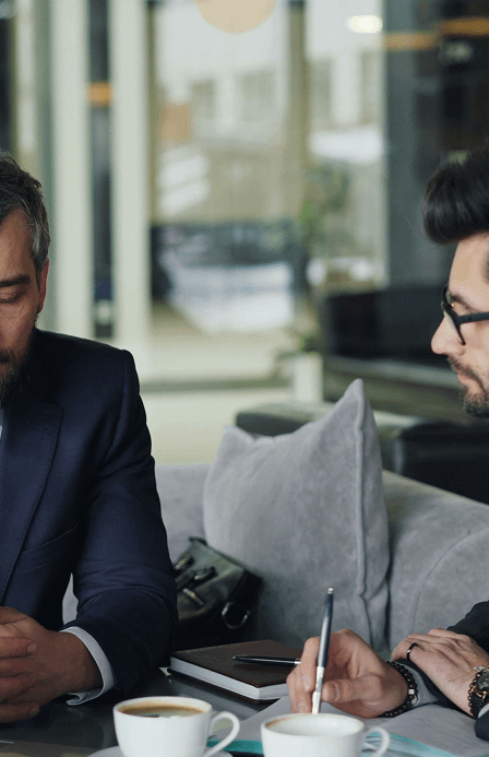 Two men in suits talking across a table with coffee cups, one taking notes